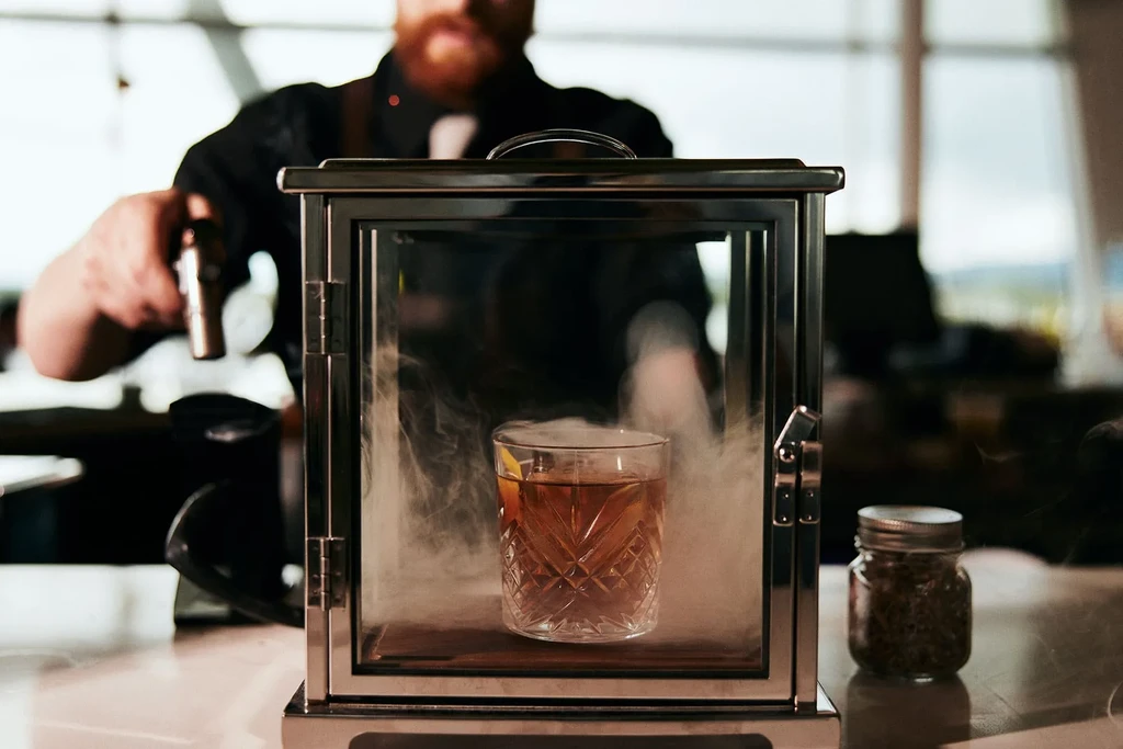 Smoked cocktail in a glass cloche, prepared by a bartender.