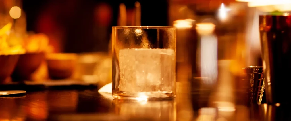 Glass of ice on a bar, with warm, blurred background lights.