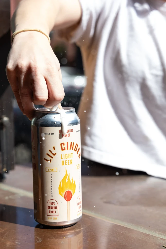 Hand opening 'LIL' CINDER LIGHT BEER' can, beer foam splashing out.