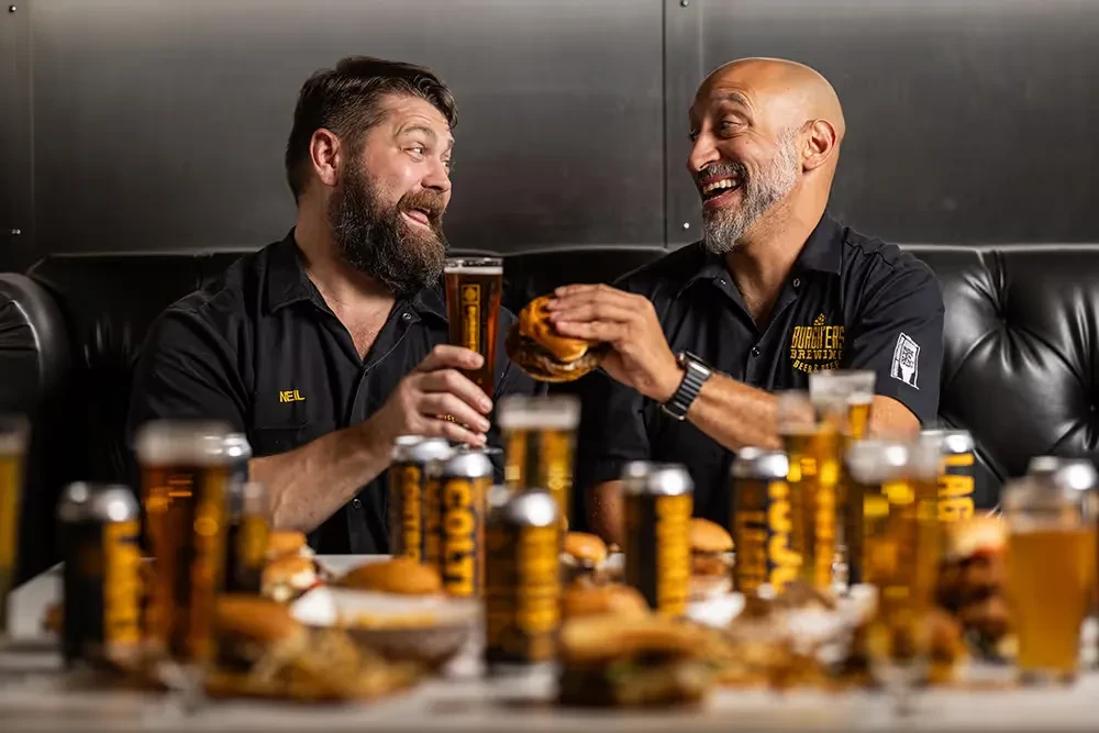 Neil and a bald man laugh over beer and burgers.