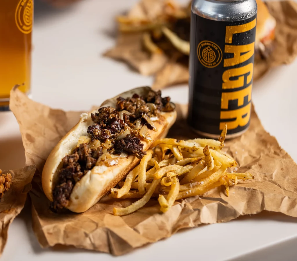 Cheesesteak sandwich, fries, and Burgher's Brewing Lager beer on paper.