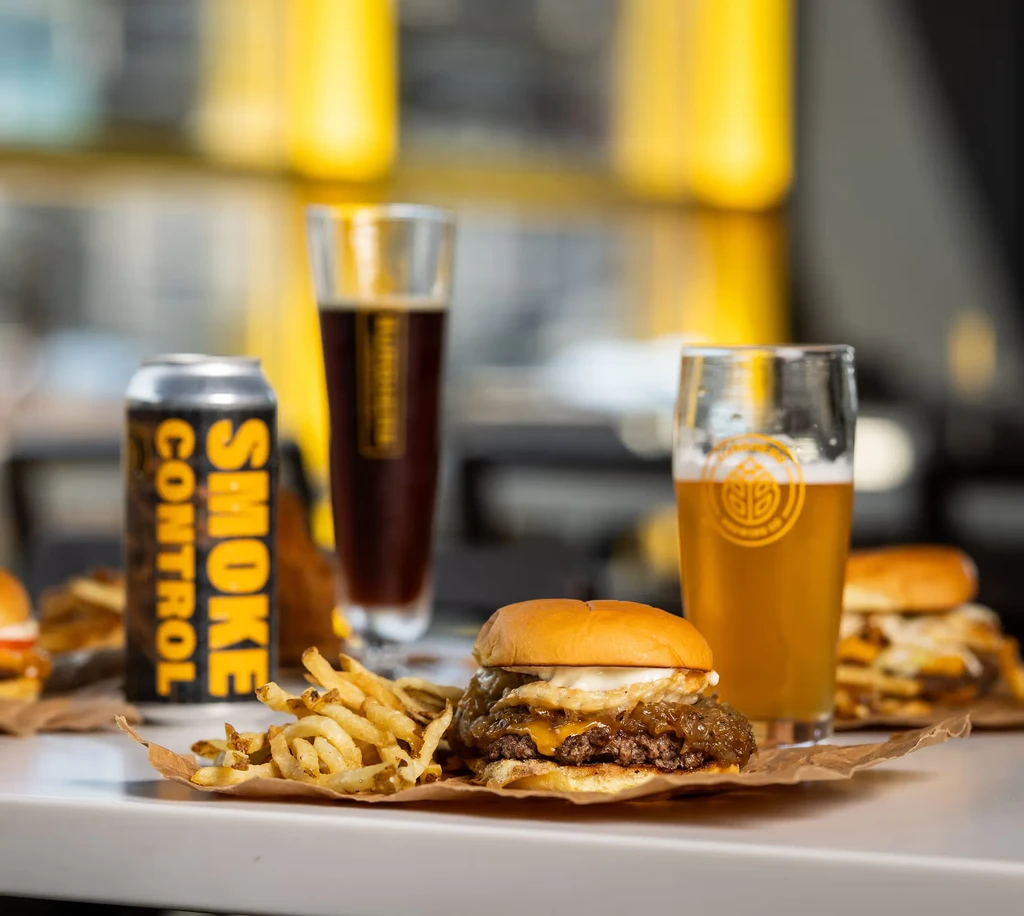 Burger with egg, fries, black beer can, and two glasses of beer.