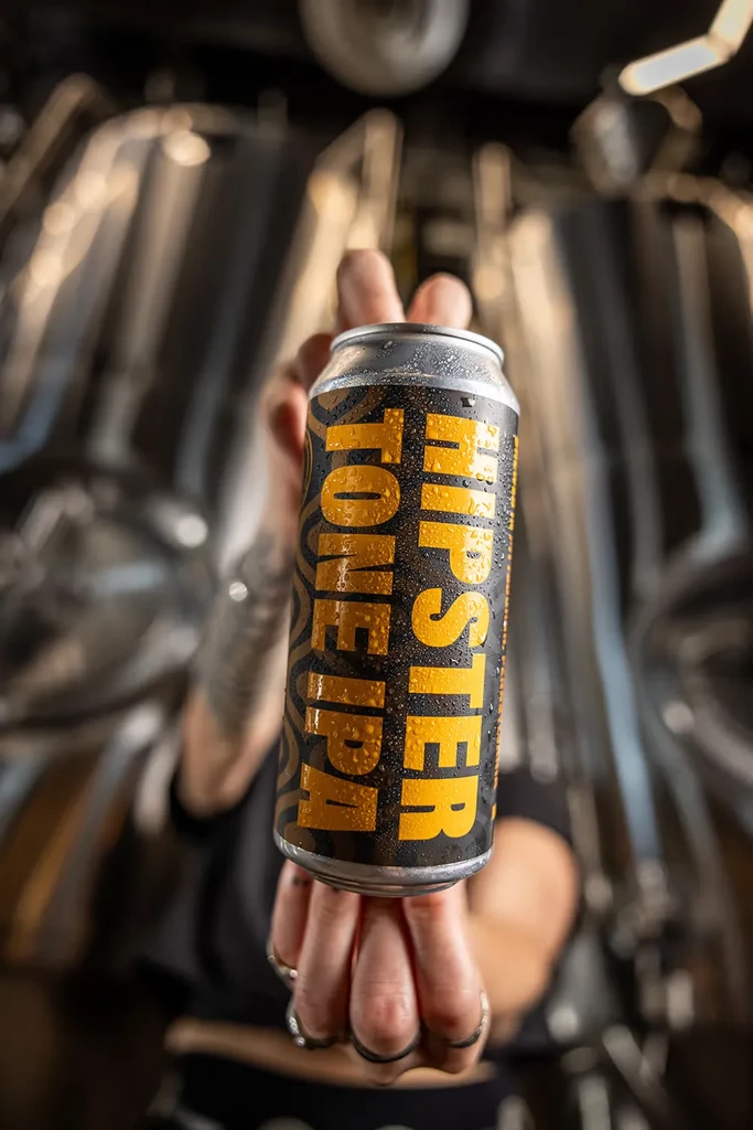 Person holds a wet black 'HIPSTER TONE IPA' can in a brewery.