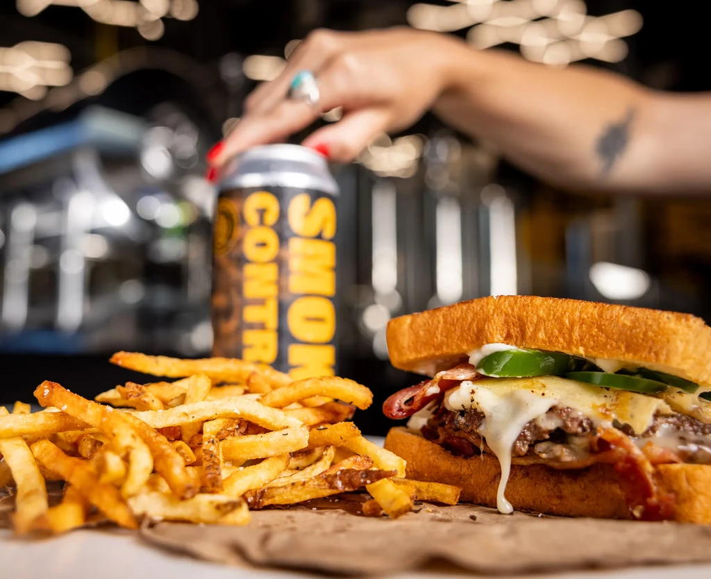 Gourmet sandwich, seasoned fries, and hand with ring by beverage.