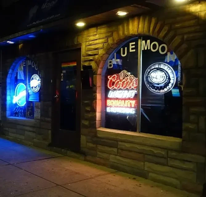 Nighttime stone bar facade with colorful neon signs and a rainbow flag.