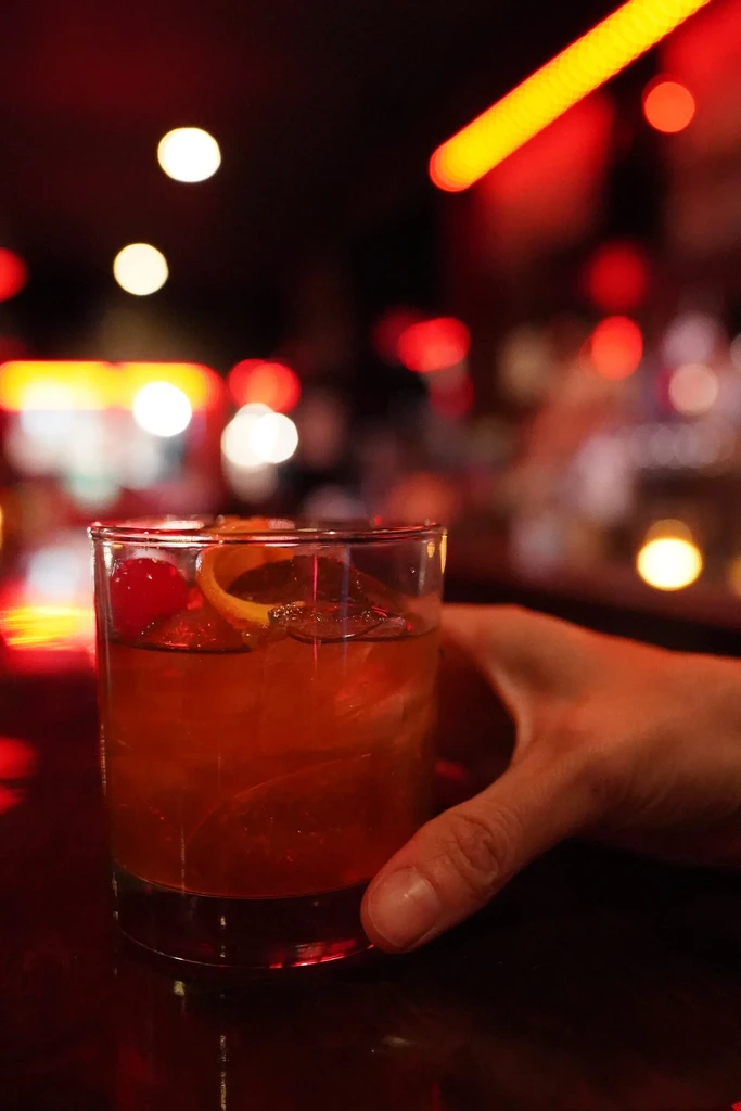 Hand holds amber cocktail with cherry and orange peel in blurred bar.
