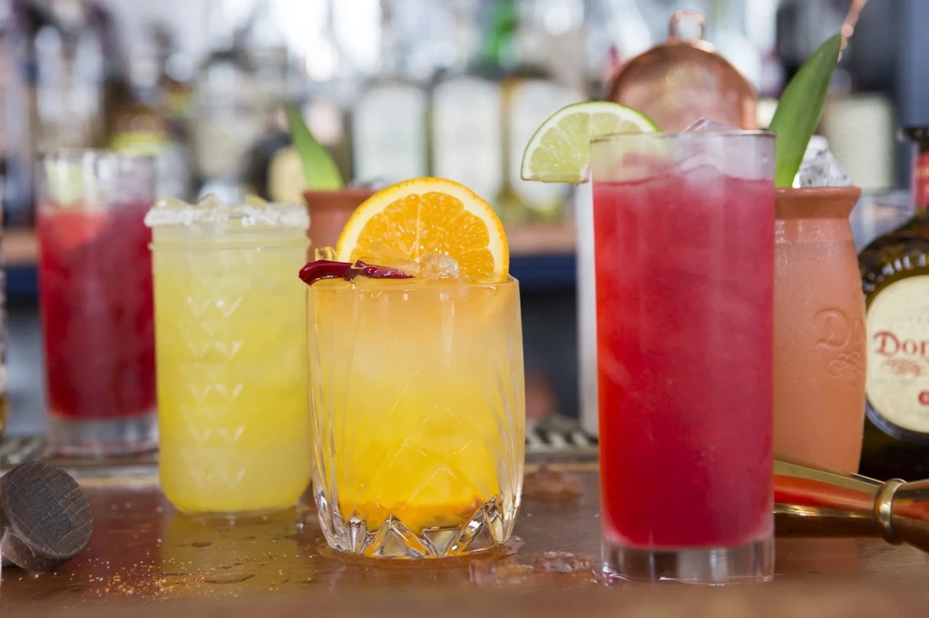 Five colorful cocktails with garnishes arranged on a bar counter.