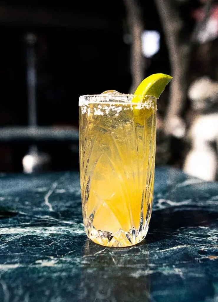 Yellow-orange cocktail in ornate glass with salted rim and lime.
