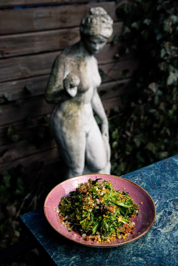 Vibrant salad in purple bowl, blurred classical statue in background.