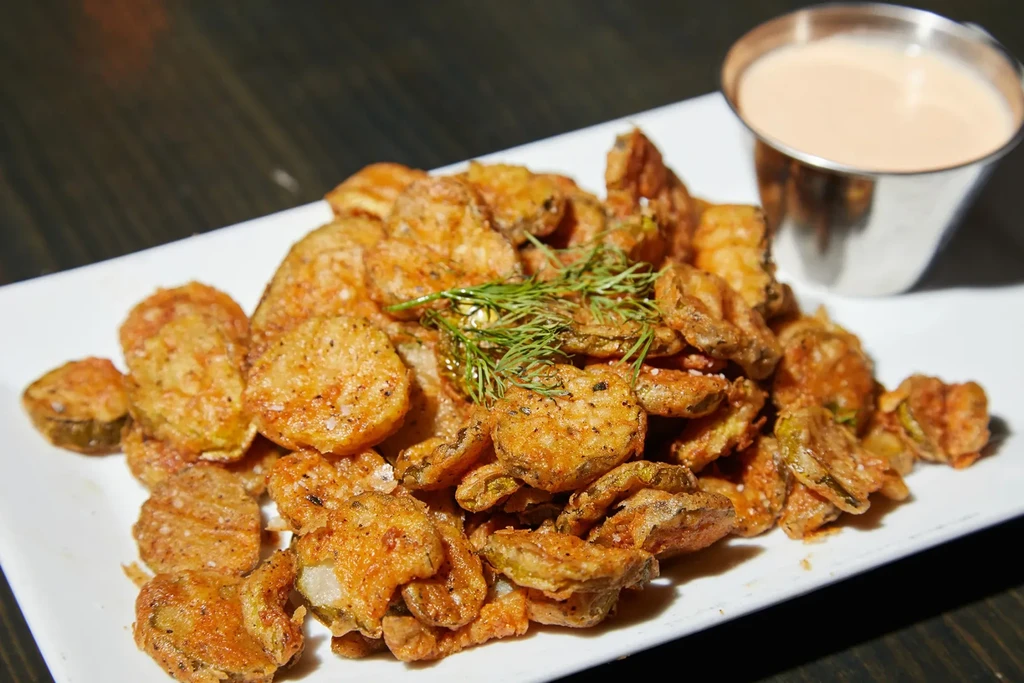Plate of golden-brown fried pickles with dill garnish and dipping sauce.