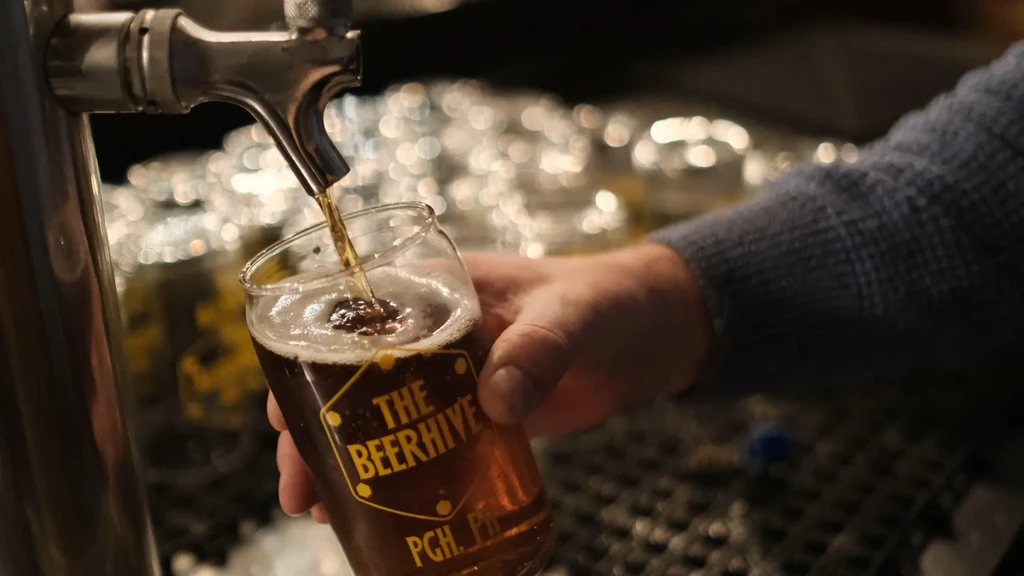Amber beer pouring into 'THE BEERHIVE' glass from a tap.
