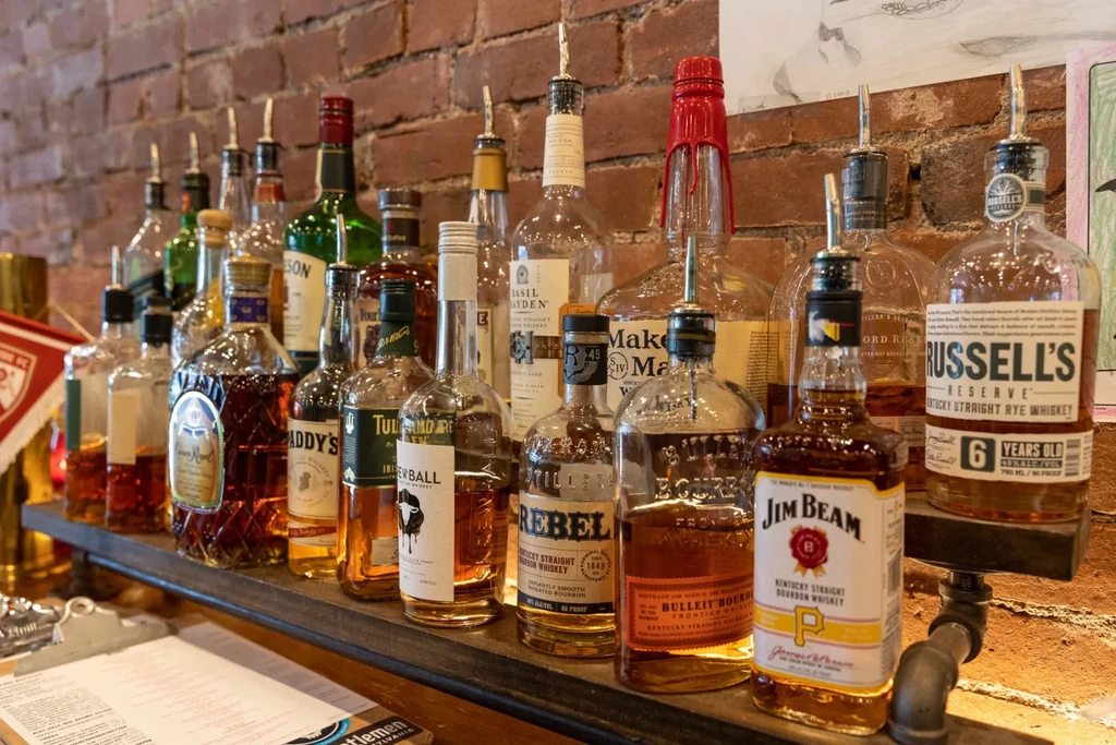 Well-stocked bar shelf with whiskey bottles, pour spouts, and a brick wall.