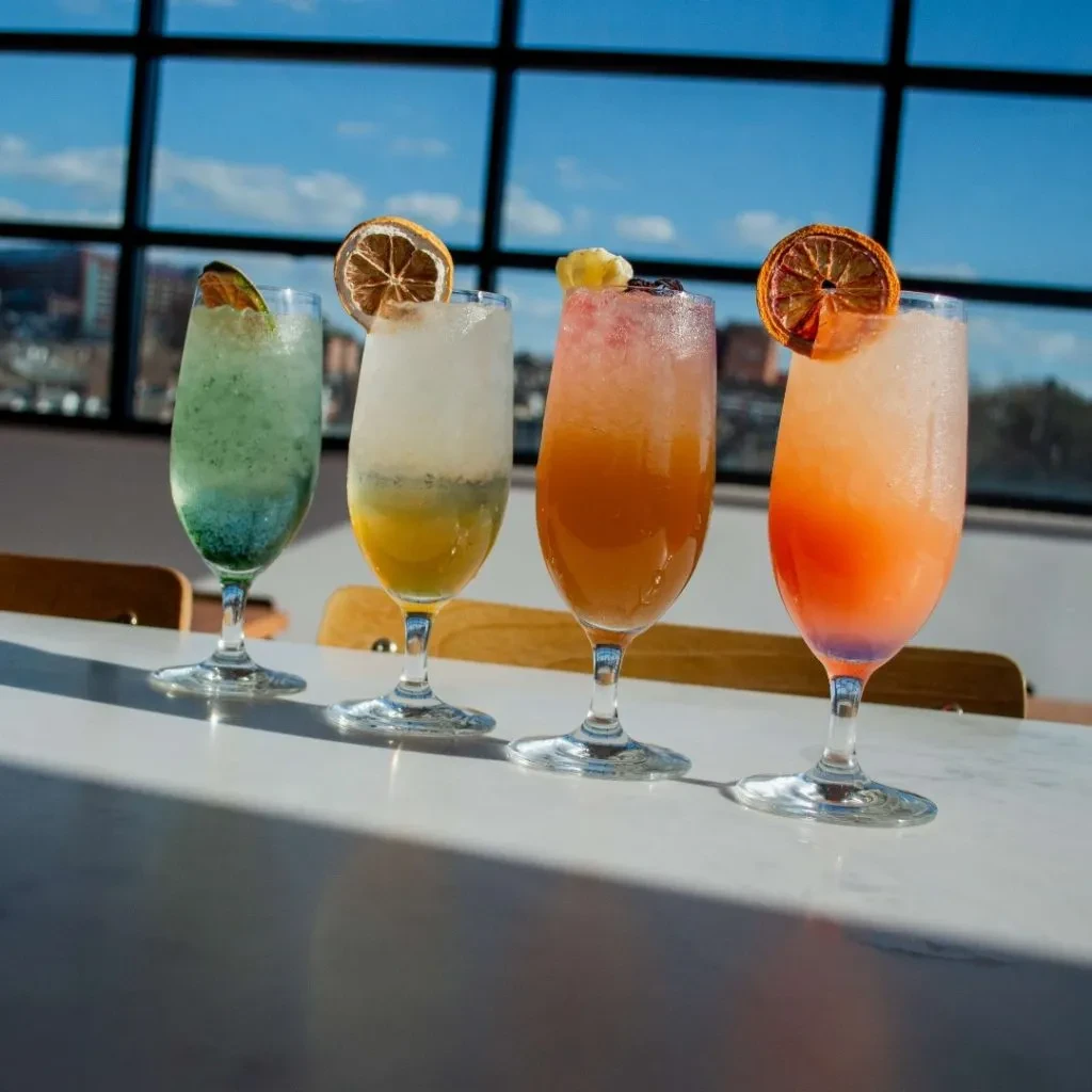 Four vibrant, layered cocktails in stemmed glasses with various garnishes.