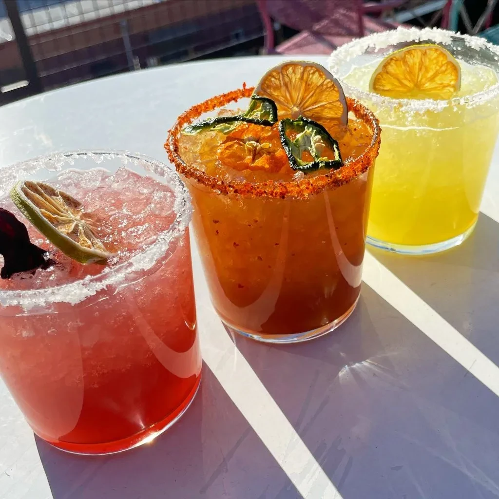 Three colorful, garnished margaritas with ice on a sunny outdoor table.