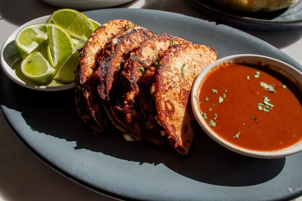 Golden-brown birria tacos, lime wedges, and dipping consommé.