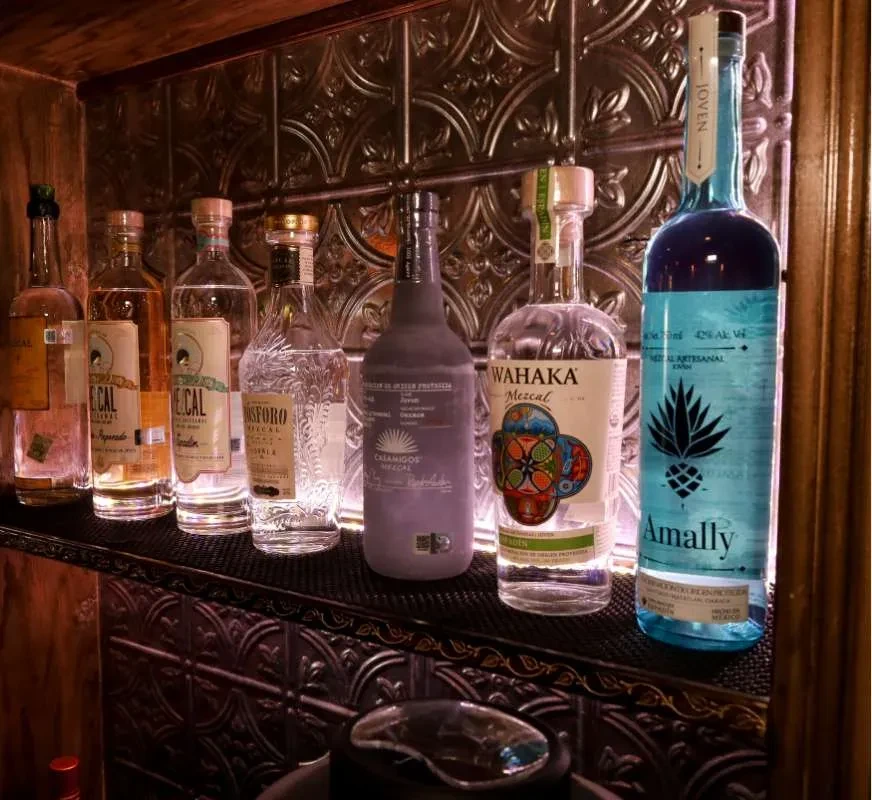 Eight mezcal and tequila bottles on a dark shelf with patterned tin wall.