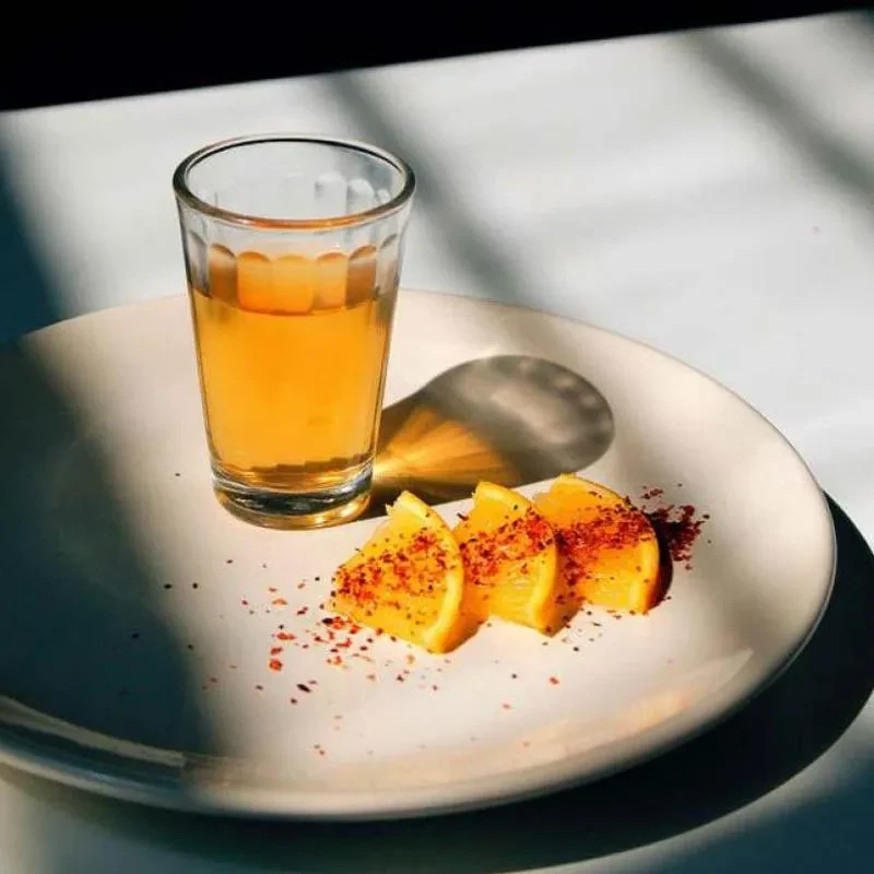 Close-up of amber drink shot with chili-dusted orange wedges on a plate.