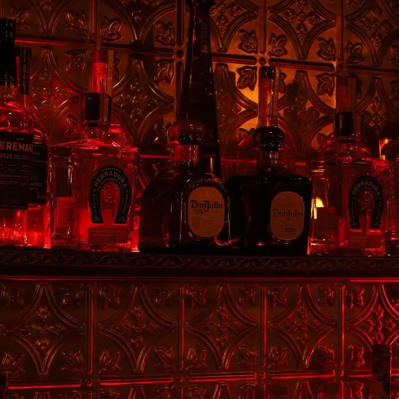 Red-lit tequila bottles on a shelf against an ornate metallic wall.