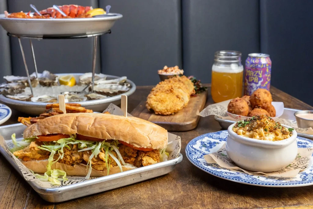 Rustic table with po'boy sandwich, fried seafood, macaroni, and various drinks.