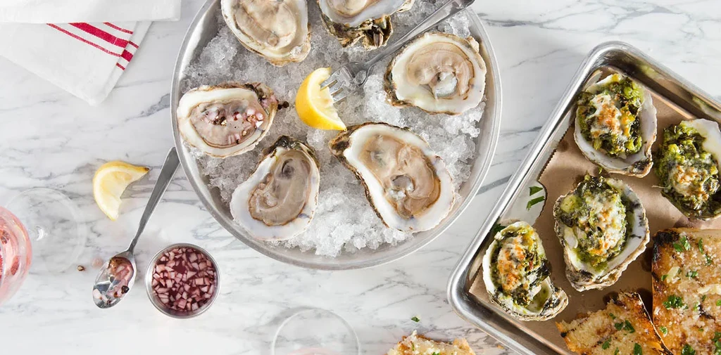 Overhead view of raw and baked oysters with sides on white marble.