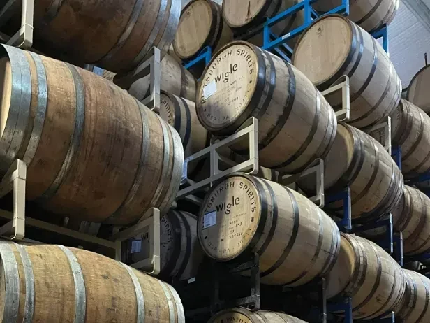 Wooden barrels labeled "Wigle Pittsburgh Spirits" stacked high on metal racks.