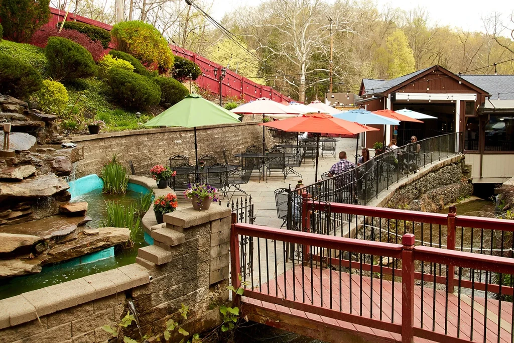 Restaurant patio with waterfall, bridge, colorful umbrellas, and green hillside.