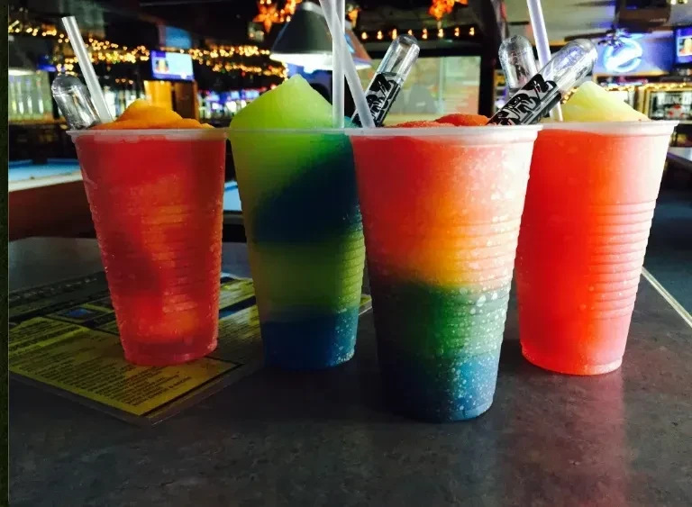 Four colorful frozen drinks with straws and tubes on a bar.