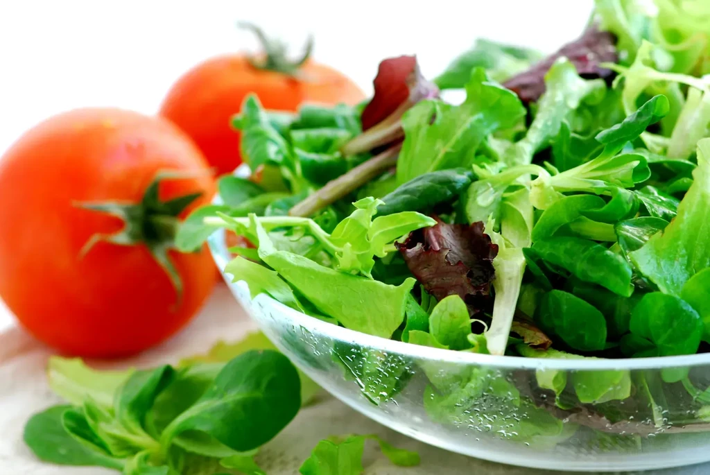 Fresh mixed green salad in glass bowl with blurred red tomatoes.