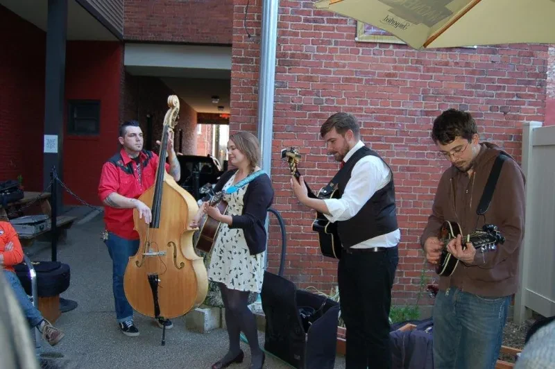 Four musicians play bass, two guitars, and mandolin outdoors.
