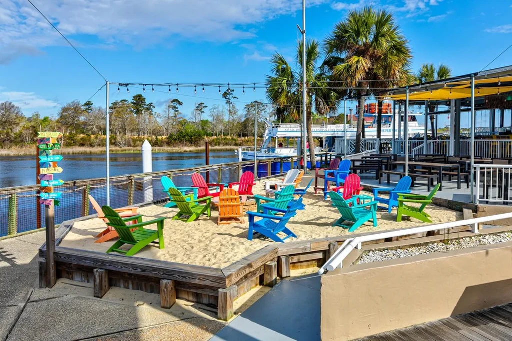 Outdoor waterfront restaurant with colorful Adirondack chairs in sand.
