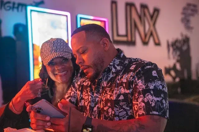 Man and woman smiling, looking at a phone in a neon-lit bar.