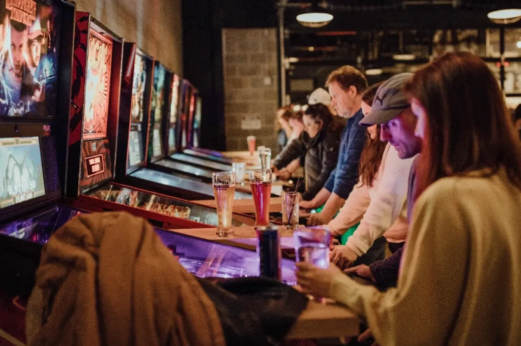 People playing brightly lit pinball machines with drinks nearby.
