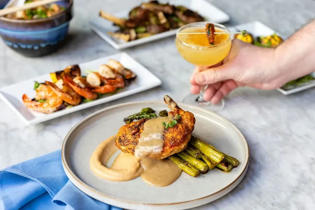 Hand holding cocktail near roasted chicken, asparagus, shrimp, and lamb.