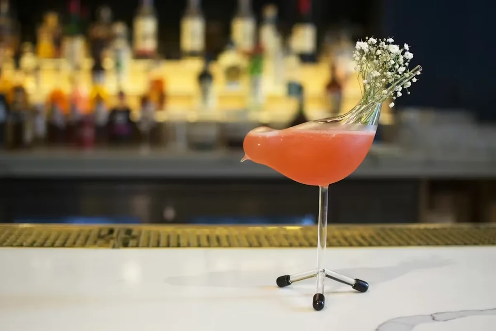 Pink cocktail in a bird-shaped glass garnished with baby's breath.