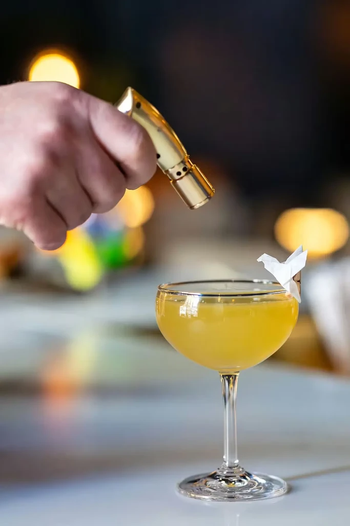 Flaming yellow cocktail in coupe glass with a white origami bird.