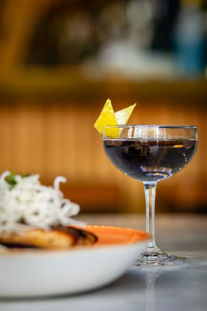 Dark cocktail with lemon peels in coupe glass, with blurred food.