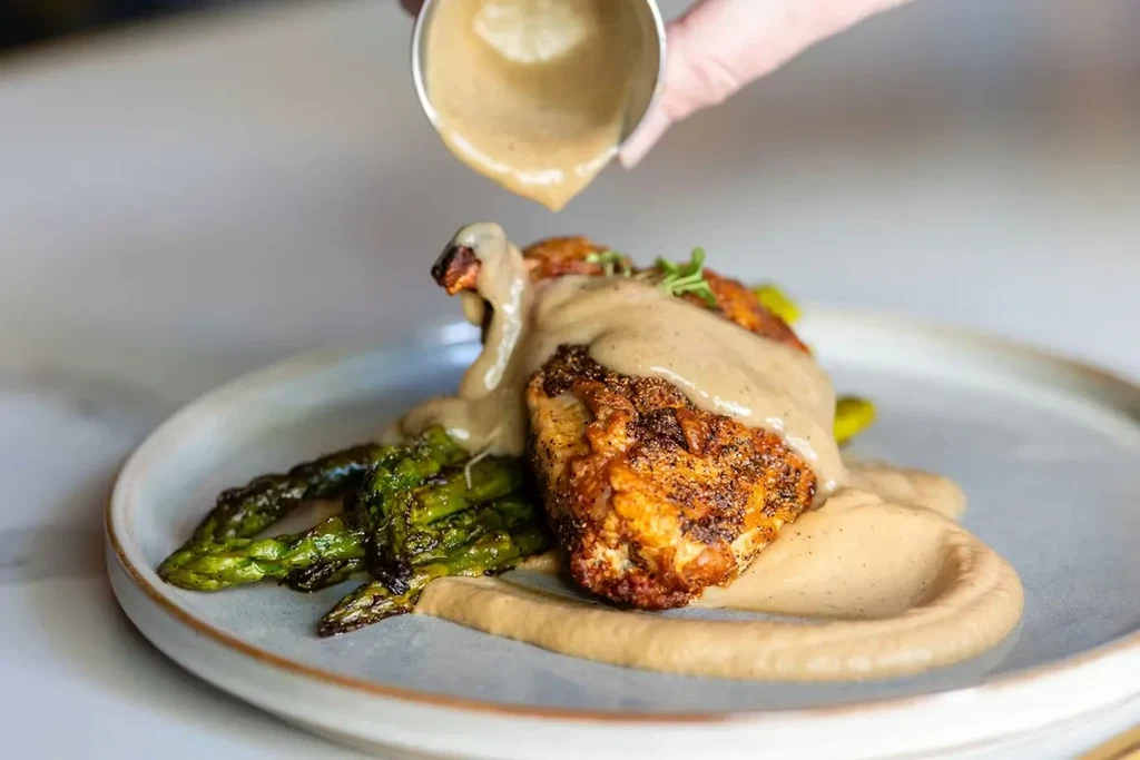 Hand pouring sauce over plated seared food and grilled asparagus.