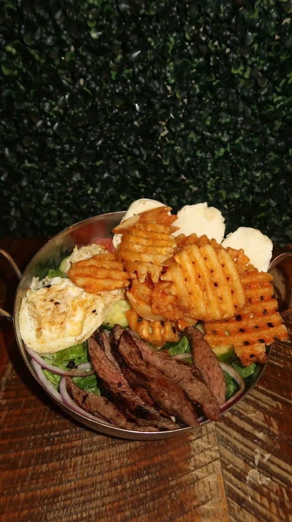 Grilled steak salad with fries and eggs in a bronze bowl on wood.