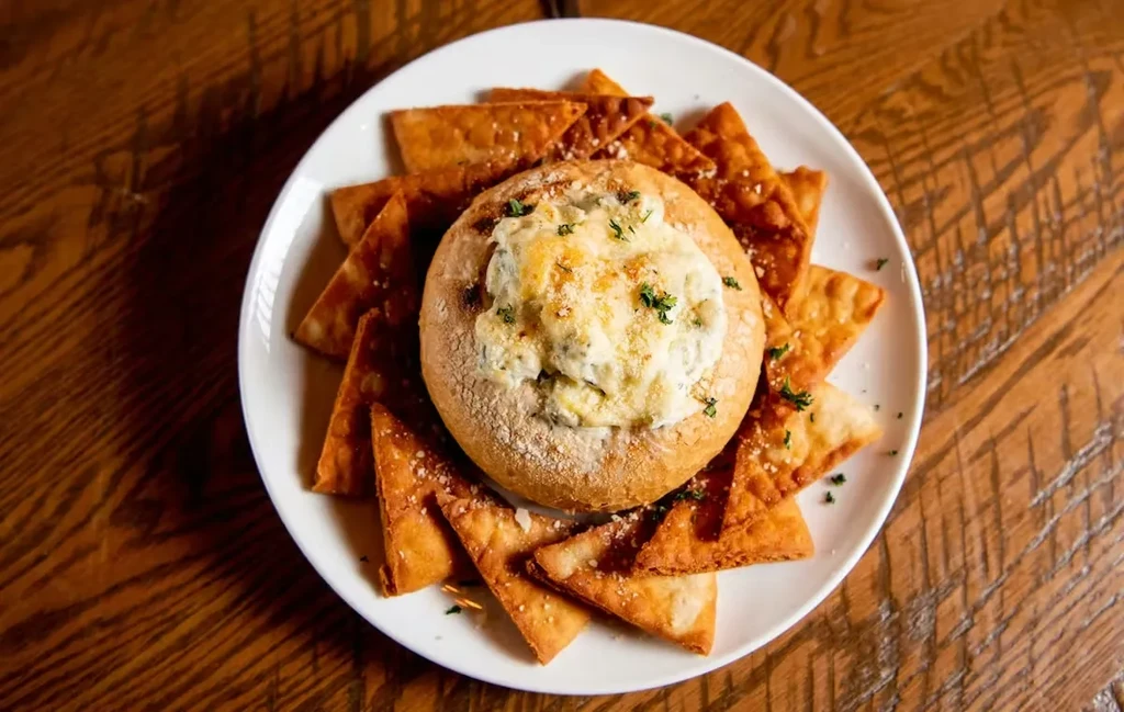Bread bowl of cheesy dip, melted cheese, herbs, chips, wood table.