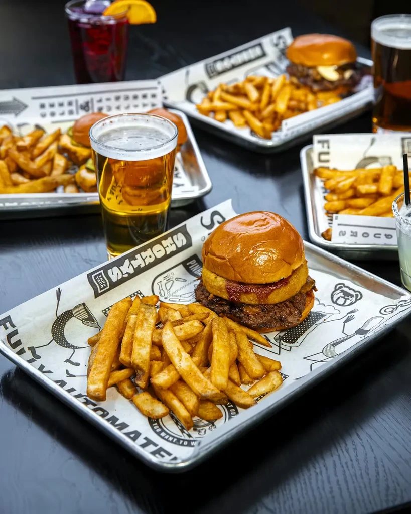 Close-up of burgers, fries, beer, and cocktails on a dark table.