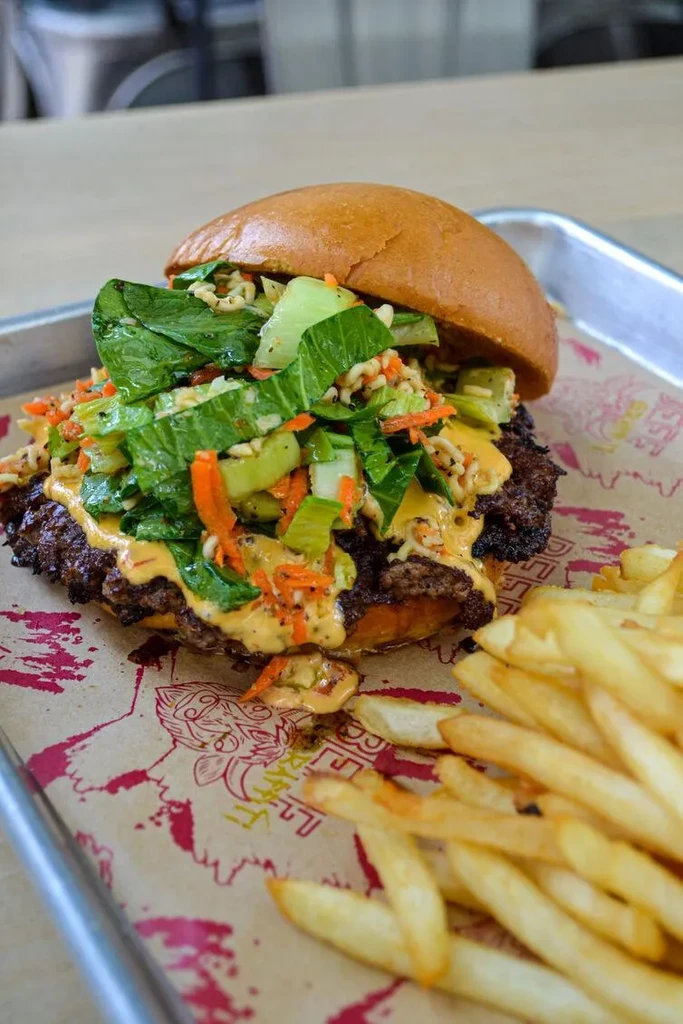 Gourmet burger with crispy ramen, vegetables, and fries on a tray.