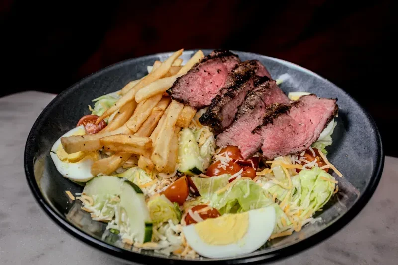 Medium-rare steak, french fries, and salad with eggs in a dark bowl.