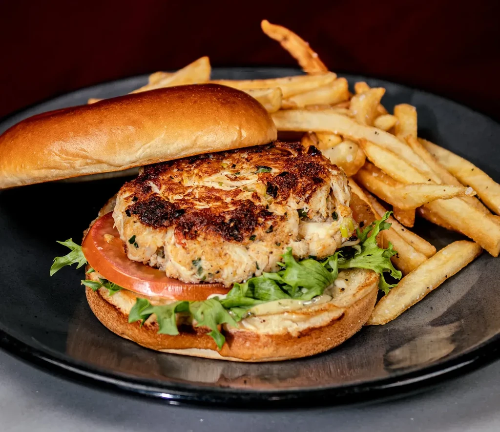 Close-up of a crab cake sandwich with french fries on a dark plate.