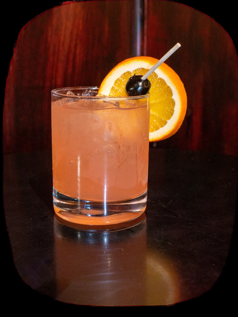Peach-colored cocktail with ice, orange slice, and cherry garnish.