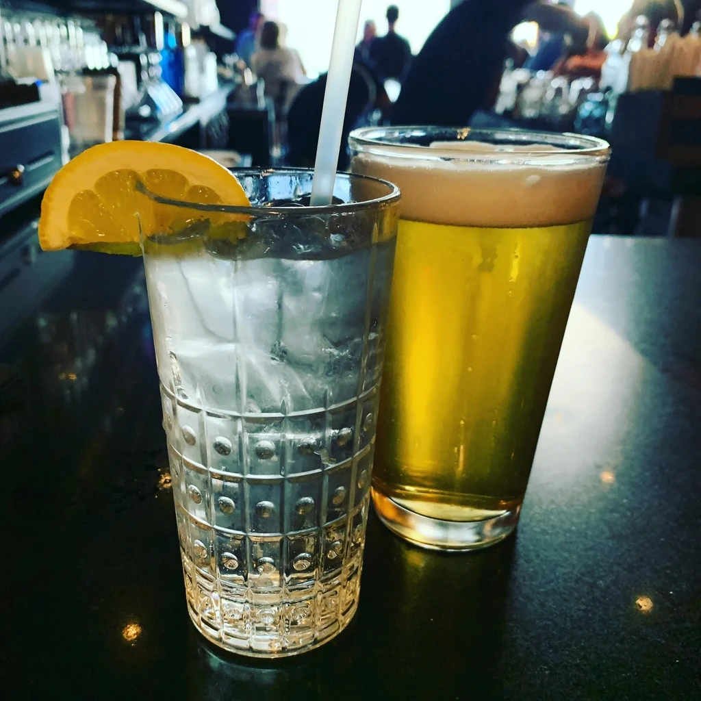 A clear drink with lemon and a golden beer on a dark bar.