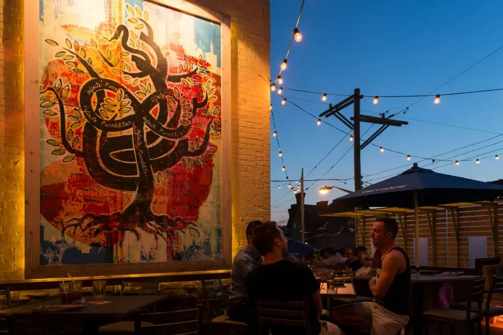 Vibrant patio with illuminated mural, string lights, and seated patrons at dusk.