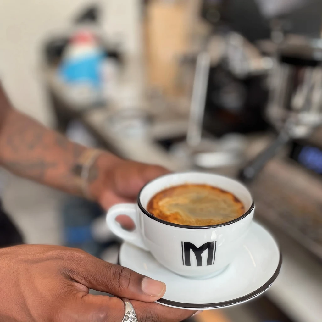 Dark hands hold 'M' coffee cup with frothy crema in a cafe.
