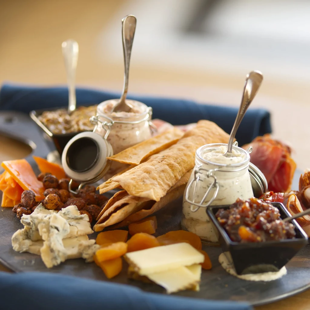Appetizer platter with cheeses, nuts, fruit, crackers, dips, and cured meat.