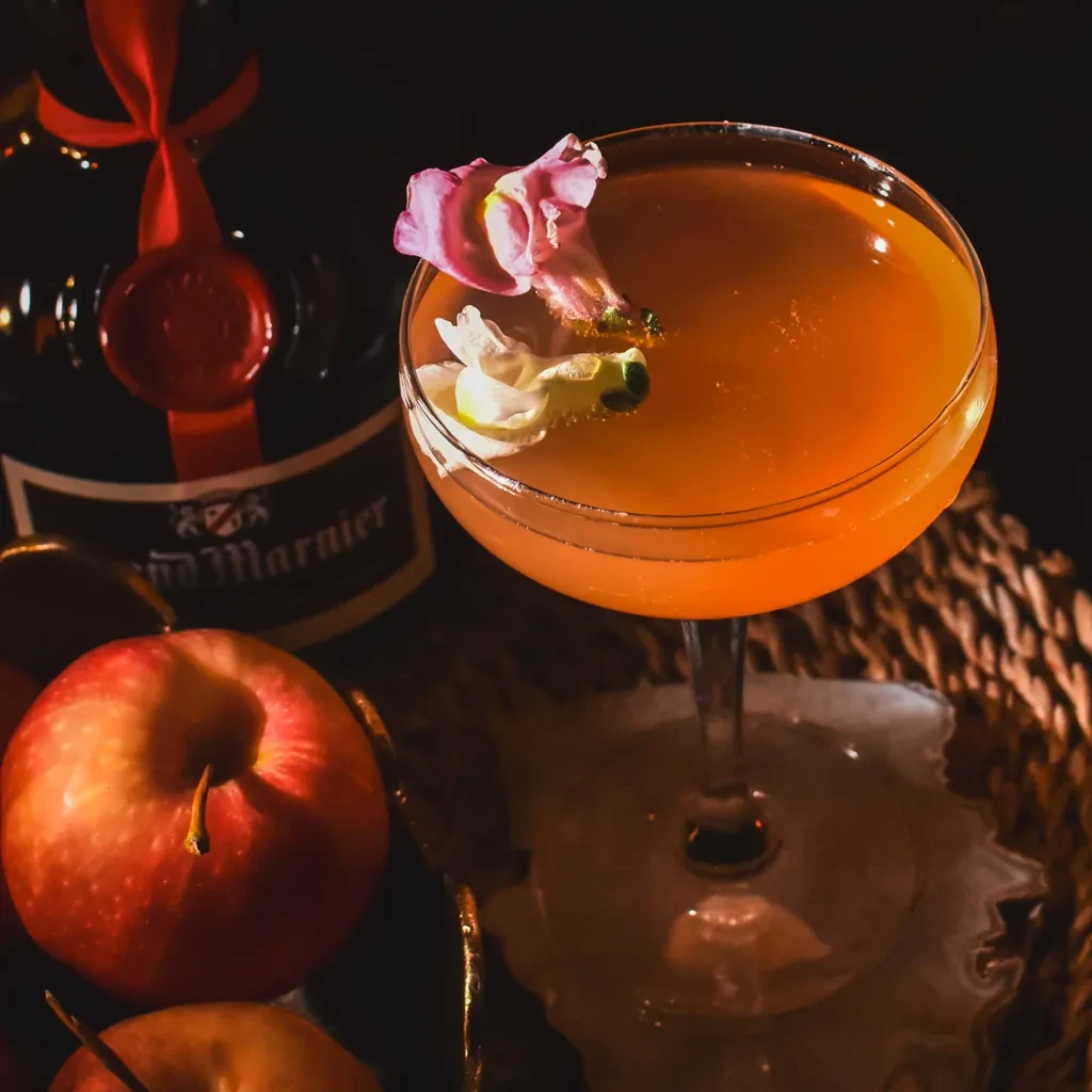 Amber cocktail with flower in coupe glass, Grand Marnier, and apples.
