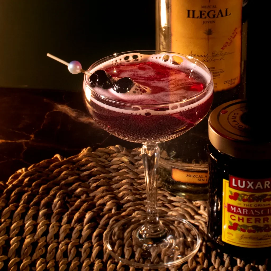 Reddish-purple cocktail in coupe, frothy, with cherries, Mezcal and Luxardo bottles.